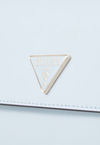 Superficie in pelle azzurro chiaro con una placca triangolare del logo Guess U.S.A. bordata in oro, caratterizzata da un simbolo di punto interrogativo.