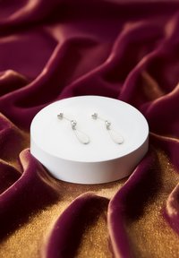 Orecchini a goccia in argento con attacco a sfera e forma a goccia, esposti su un piedistallo rotondo bianco contro un tessuto testurizzato.