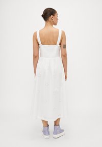 Hvit midi-kjole med justerbare stropper, teksturert liv, og broderte blomsterdetaljer. Brukt med lyse lilla høye sneakers.