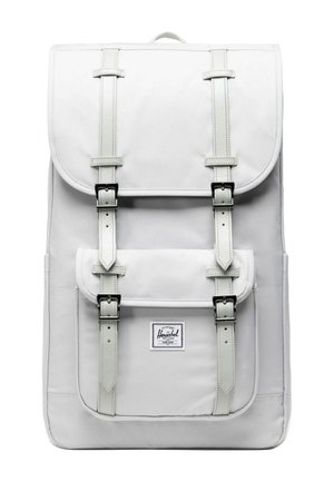 Sac à dos rectangulaire blanc avec deux rabats fermés par des sangles beige et des boucles en métal noir, présentant une poche avant avec une étiquette de marque.