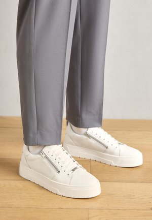 Sneakers bianchi con finitura testurizzata, dotati di una cerniera laterale, un davanti con lacci e una spessa suola bianca. Abbinati a pantaloni grigi.