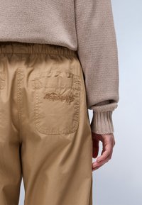 Beige, lette bukser med elastisk midje, med to baklommer og broderte detaljer. Strikkegenser med ribbestrikkede mansjetter.