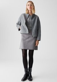Giacca grigia con texture e colletto rialzato, indossata sopra una camicia grigia a bottoni, abbinata a una gonna in suede grigia e stivaletti neri alla caviglia.
