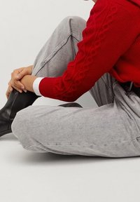Roter Strickpullover mit Zopf-Muster, kombiniert mit grauen weiten Hosen und schwarzen Ankle-Boots, in Sitzposition vor einem neutralen Hintergrund.