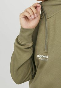 Olivgrön kvartslång ziptröja med en mjuk textur, som har en dragkedja och en vit textlogga på bröstet.