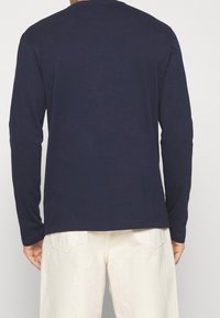 Man som bär en marinblå långärmad skjorta och off-white byxor, sedd bakifrån med händerna avslappnat vid sidorna.