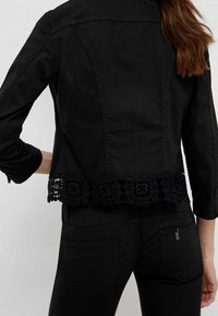 Mujer de espaldas vistiendo una chaqueta negra ajustada con borde de encaje floral y jeans negros, con el cabello castaño sobre el hombro.