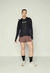 Woman standing wearing black zip-up jacket, black t-shirt, brown shorts, black socks, and white sneakers against plain background.