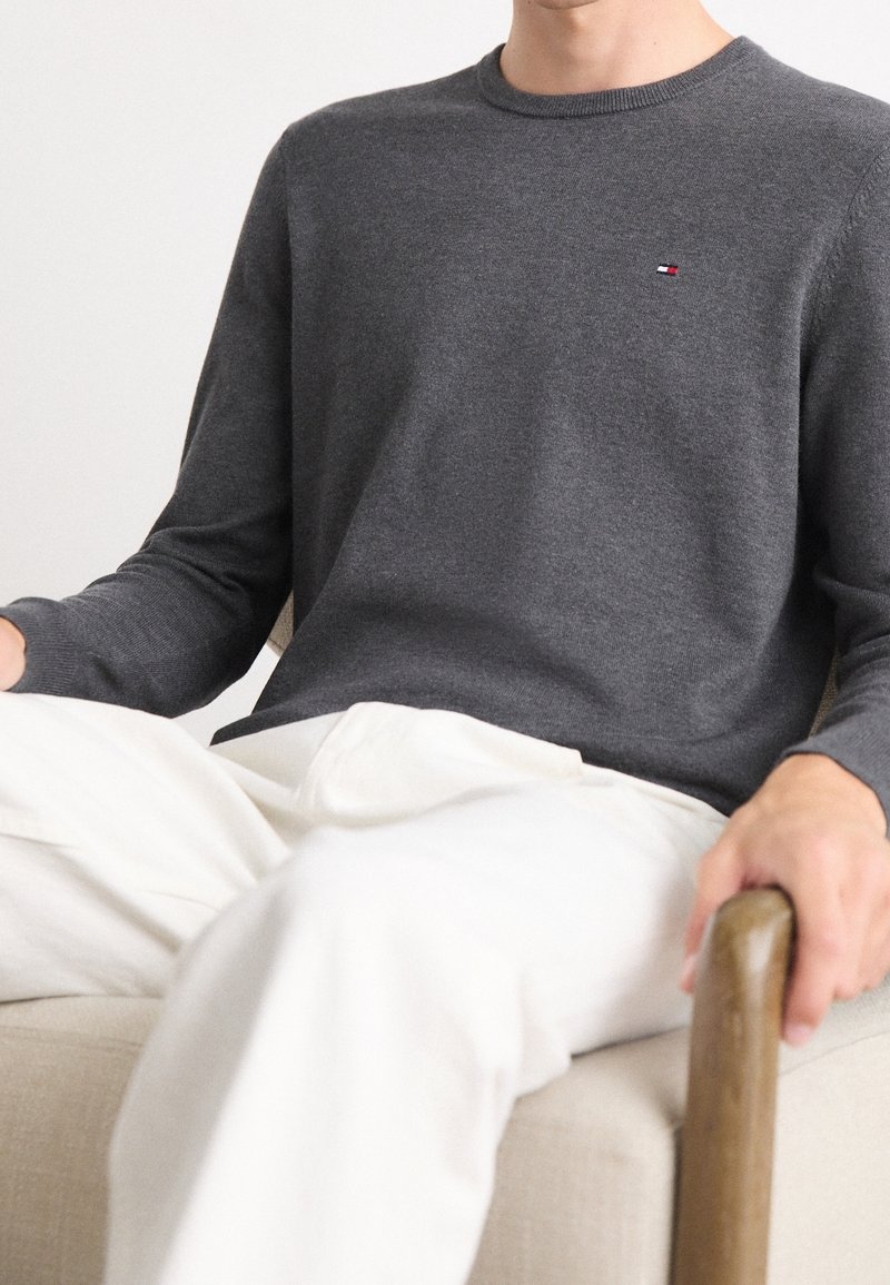 Homme assis sur une chaise beige portant une chemise à manches longues gris foncé et un pantalon blanc, la main reposant sur un accoudoir en bois.