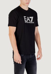 Camiseta de algodón negra con cuello redondo que presenta un destacado logo blanco de EA7 y el texto 'EMPORIO ARMANI'. Diseño sencillo, con mangas cortas.