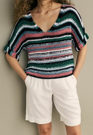 Pull tricoté multicolore avec un col en V, présentant des rayures vertes, roses, blanches et noires. Associé à un short en lin blanc.