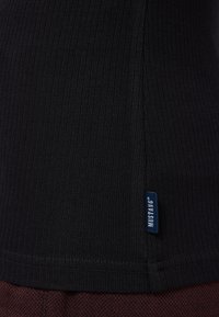 Czarny prążkowany top na ramiączkach o dopasowanym kroju z małą niebieską metką z napisem "MUSTANG" przy krawędzi, prezentujący teksturowaną tkaninę.