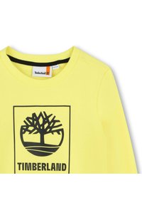Sweatshirt jaune avec un logo Timberland noir arborant un design d'arbre et le mot "TIMBERLAND" en dessous. Encolure ronde côtelée noire.