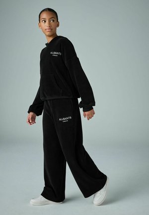Sort velour træningsdragt med en hættebluse med en frontlomme og bredde bensnit. Begge dele har "ALLSAINTS LONDON" branding. Hvide sneakers.