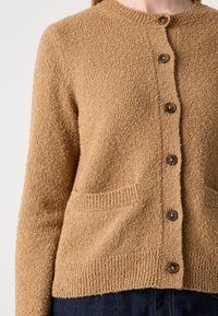 Primo piano di un cardigan a maglia marrone con una texture, maniche lunghe, scollatura rotonda, bottoni frontali e tasche e orlo leggermente a coste.