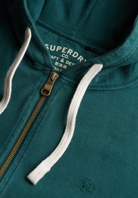 Sudadera con cremallera en color verde azulado, con cordones blancos, tirador de cremallera de latón etiquetado como "Superdry" y etiqueta de marca interna parcialmente visible.