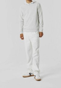 Person wearing light gray hoodie, white pants, and white sneakers with black stripes, standing against a plain white background.