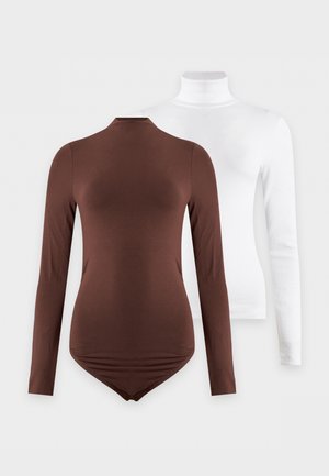 Femme aux cheveux bouclés portant un col roulé marron ajusté, un pantalon noir taille haute et des chaussures noires, debout devant un fond blanc uni.