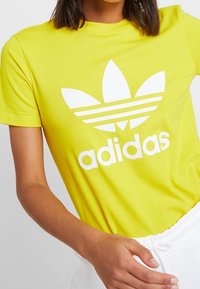 Gul bomullströja med en vit Adidas-logotyp och den ikoniska tre-randsdesignen över bröstet; standard rundad halsringning.