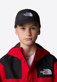 Gorra negra con un logo blanco, combinada con una chaqueta roja y negra que presenta grandes bloques de color y detalles de hardware visibles. Camiseta blanca debajo.