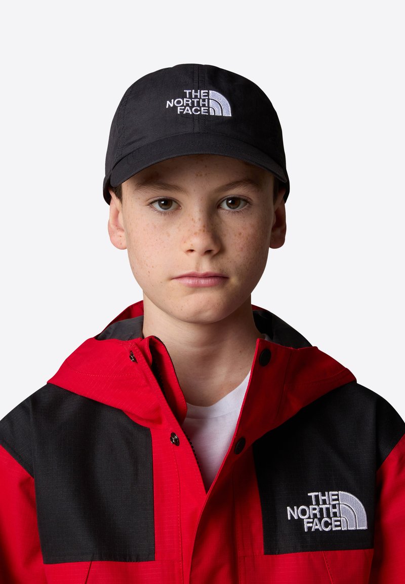 Gorra negra con un logo blanco, combinada con una chaqueta roja y negra que presenta grandes bloques de color y detalles de hardware visibles. Camiseta blanca debajo.