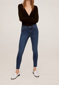 Jeans attillati di denim blu scuro, con vita alta, tasche anteriori e posteriori, e orlo sfrangiato. Accompagnati da scarpe slip-on bianche.