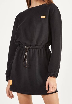 Robe noire à manches longues avec taille élastique et cordon, patches orange sur la poitrine et près de l'ourlet, portée par une personne debout.