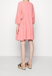 Rosa Baumwoll-Minikleid mit langen Ärmeln, gestuftem Design und strukturiertem Finish, kombiniert mit schwarzen Keilsandaletten vor einem hellen Hintergrund.
