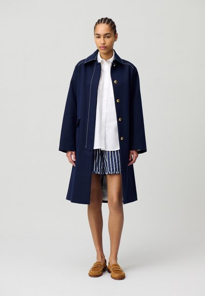Manteau bleu marine avec des boutons dorés, des détails de couture blanche et une coupe mi-longue, porté sur une chemise blanche et un short rayé.
