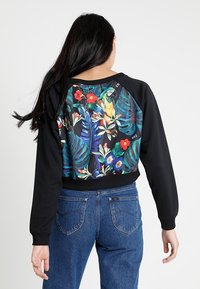 Svart sweatshirt med ett livligt tropiskt tryck med blommor och en papegoja på ryggen. Ribbad mudd vid ärmar och midja. Blend av bomullsmaterial.