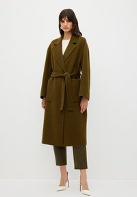 Olivgrön ylleblandad kappa med bälte i midjan, slagkrage, två framfickor och långa ärmar. Matchad med gröna byxor och vita klackar.