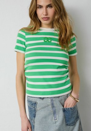 Fiatal nő zöld-fehér csíkos rövid ujjú ingben és világos farmer szoknyában, amelyen foltzsebek találhatók, egyik kezét a zsebébe dugva pózol.