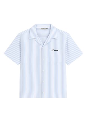 Femme portant une chemise à manches courtes rayée bleu clair avec boutons et une casquette de baseball beige avec logo, debout sur un fond uni.