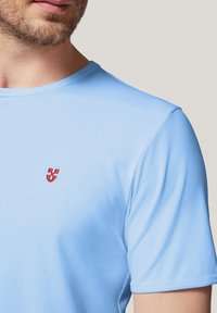 Jasnoniebieska koszulka z krótkim rękawem, wykonana z miękkiego materiału, z małym czerwonym logo na piersiach oraz okrągłym dekoltem. Gładka tekstura.