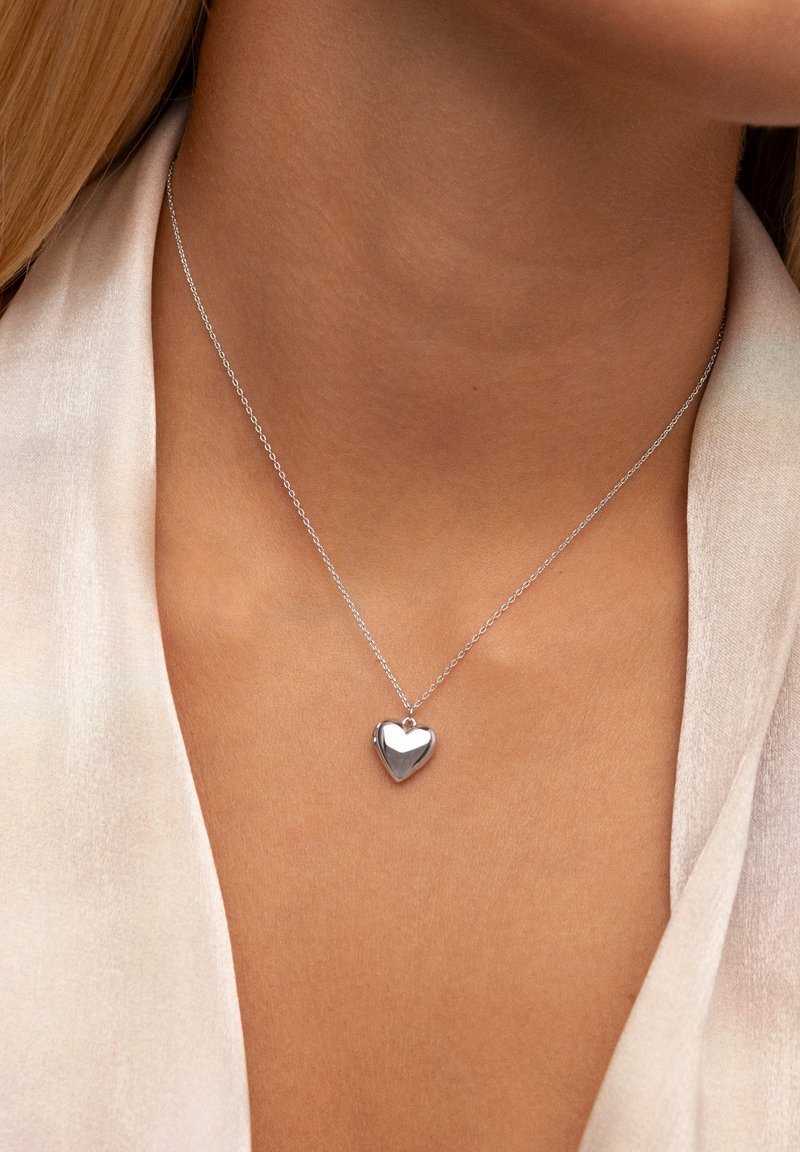 Zilveren hartvormige hanger aan een delicate ketting, met een gepolijste afwerking en een klassieke hartvorm, die rust op blote huid.