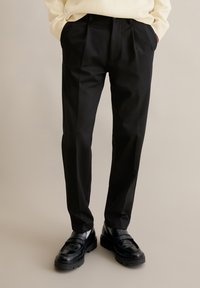 Man wearing black tailored trousers with front pleats, black leather loafers, and a cream sweater with hands in pockets.