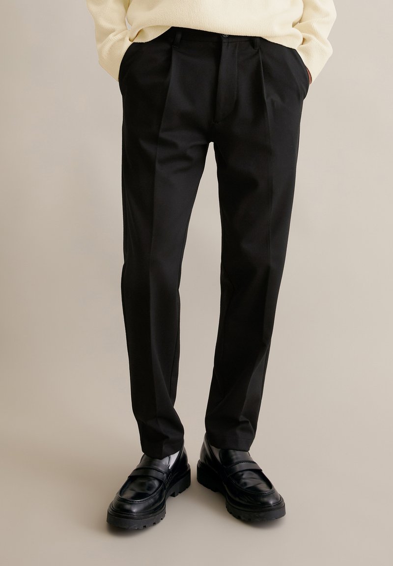 Man wearing black tailored trousers with front pleats, black leather loafers, and a cream sweater with hands in pockets.