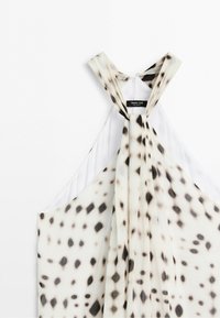 Sleeveless top with black and brown irregular spots on white fabric, featuring crossed straps and label at the back neckline.