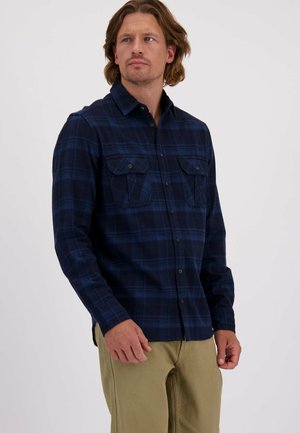 Man in een marineblauwe geruite overhemd met knopen en borstzakken, in combinatie met beige broek, die naar de zijkant kijkt tegen een effen achtergrond.