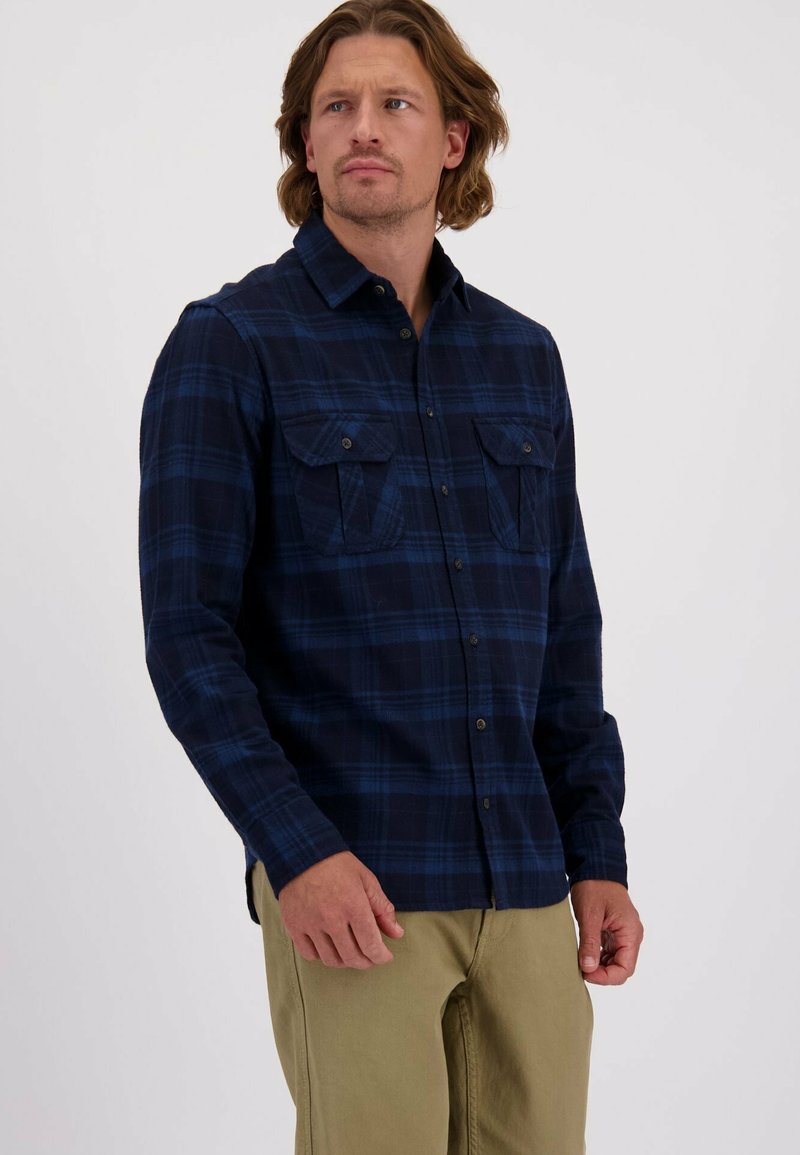 Man in een marineblauwe geruite overhemd met knopen en borstzakken, in combinatie met beige broek, die naar de zijkant kijkt tegen een effen achtergrond.