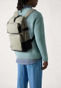Grauer Nylon-Rucksack mit zwei Fronttaschen, schwarzen Akzenten und gepolsterten Schultergurten, getragen von einer Person in einem gerippten Minzpullover und blauen Jeans.