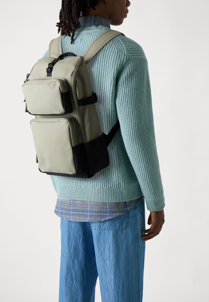 Grauer Nylon-Rucksack mit zwei Fronttaschen, schwarzen Akzenten und gepolsterten Schultergurten, getragen von einer Person in einem gerippten Minzpullover und blauen Jeans.