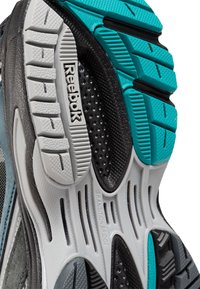 Gros plan d'une semelle de chaussure Reebok montrant des motifs de crampons texturés et des matériaux en caoutchouc bleu, noir, blanc et gris.