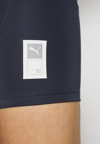 Puma sportshorts i marinblått tyg, med en fyrkantig vit logotagg med en grå Puma-logga och texten "FIRST MILE". Slät textur.