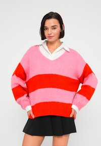 Pull surdimensionné à col en V avec des rayures horizontales roses et rouges. Texture tricotée, manches longues et poignets blancs. Associé à une jupe noire.