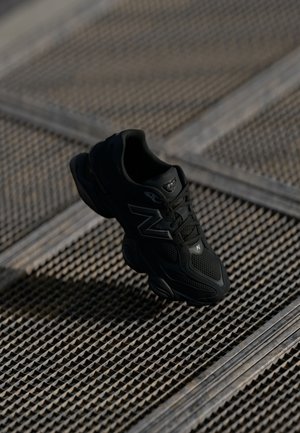 Czarny sneaker New Balance z detalami z siateczki i skóry unoszący się nad teksturowaną metalową powierzchnią w miękkim oświetleniu.