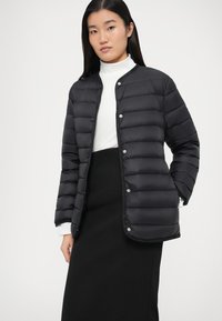 Γυναίκα που φοράει μαύρο πουπουλένιο σακάκι με κουμπιά πάνω από λευκό ζιβάγκο και μαύρη φούστα, στέκεται μπροστά σε ένα απλό ανοιχτό φόντο.