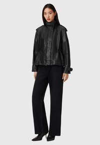 Zwarte lederen jas met hoge kraag, gestructureerde afwerking en ritssluiting aan de voorkant, gecombineerd met wijde zwarte jeans en puntige zwarte schoenen.
