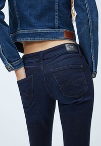 Mörkblå denimjeans med två bakfickor, synlig sömnad och en svart varumärkesetikett vid midjan, kombinerat med en matchande denimjacka.