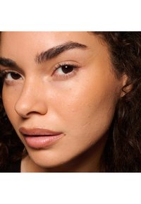 Primer plano de un rostro con tono de piel medio, cejas definidas, textura de piel suave y color natural de labios. El cabello es oscuro y rizado.